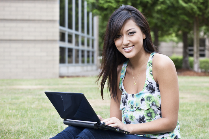 Cute Asian girl sitting in front of her school using a laptop. She's smiling at the camera.